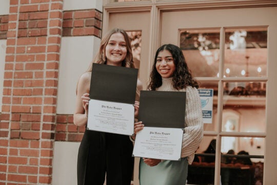 Two inductees smiling and holding membership certificates outside of Town and Gown.