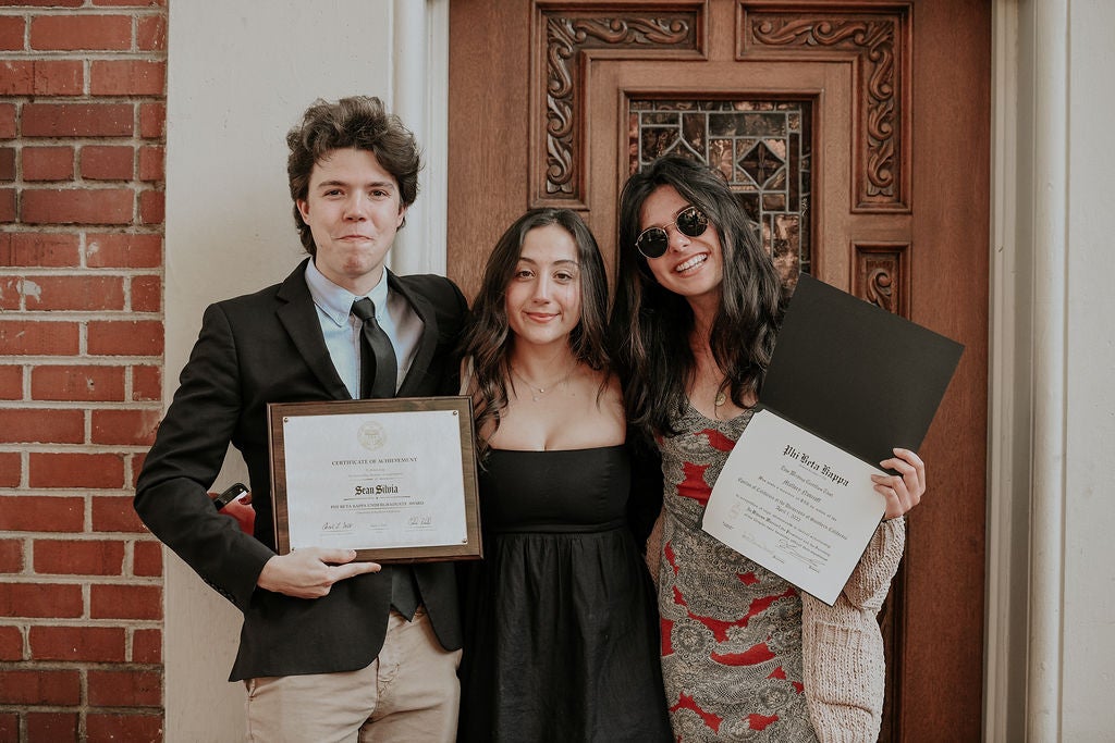 An award recipient holding plaque, a guest, and an inductee holding membership certificate, standing at doorway. All are smiling.