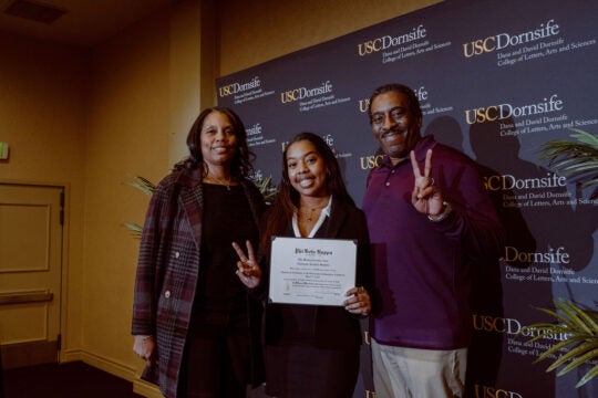 Inductee smiling while holding membership certificate. Standing with two guests. Inductee and guest have fingers held in the Trojan Victory sign.