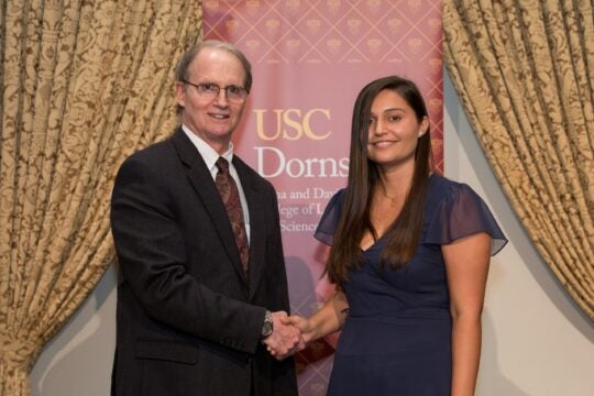 PBK Board member, Greg Thalmann, shaking hands with inductee on stage. In the background is a USC Dornsife banner between parted gold drapes.