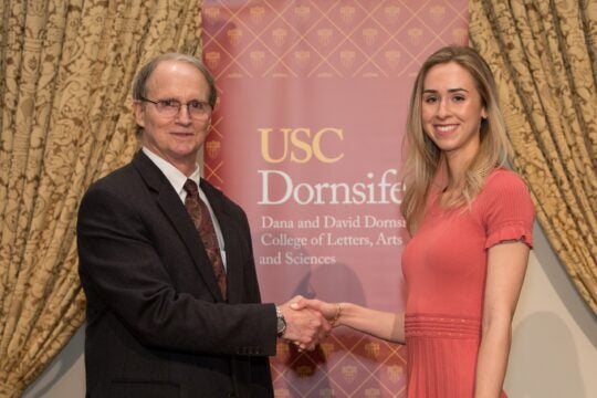 PBK Board member, Greg Thalmann, shaking hands with inductee on stage. In the background is a USC Dornsife banner between parted gold drapes.