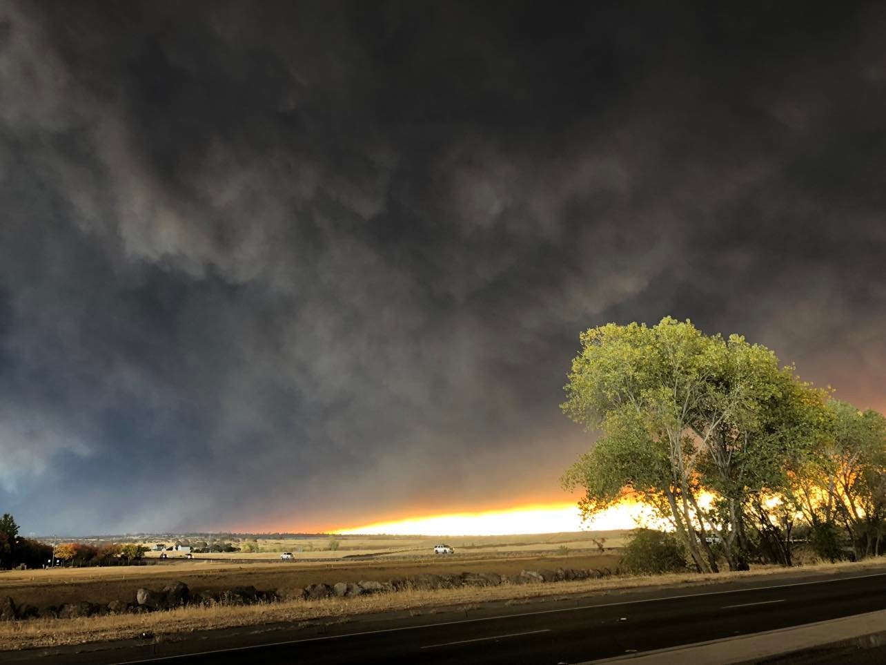 Dark smoke covers the sky of wide expanse of land with a highway in the foreground and vehicles off in the distance.  There is a bright blaze of a fire on the horizon.