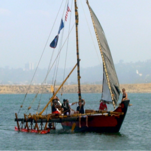 A catamaran with a sail and people aboard in a body of water with the shore in the background.