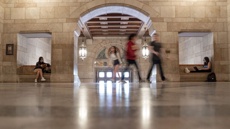 Inside of Doheny Library entry way. Two out of focus people walk by as a student on the left and a student on the right sit on benches studying.
