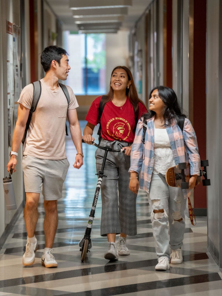 Three students talk as they walk together down a hallway in Irani Hall. One student is carry a skateboard, another is pushing a scooter and the third is holding a reusable water bottle.