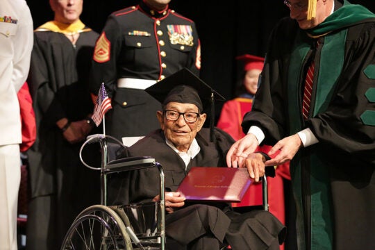 A person in a wheelchair accepts a diploma wearing their commencement cap and gown