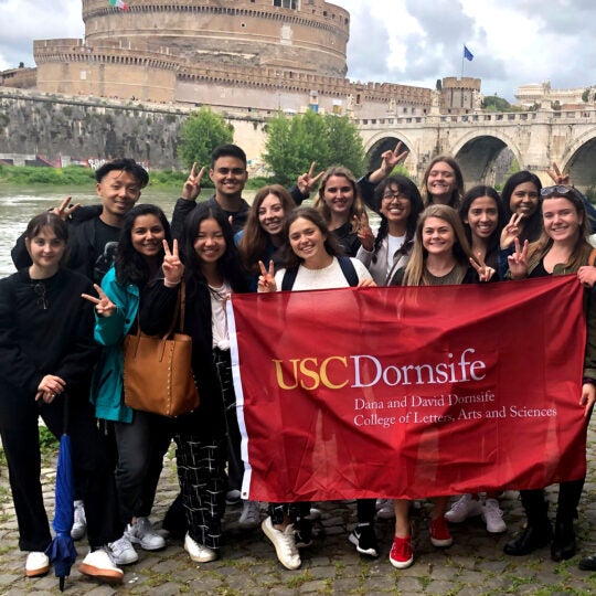 group shot of students with USC flag in Italy, ancient ruins in the background