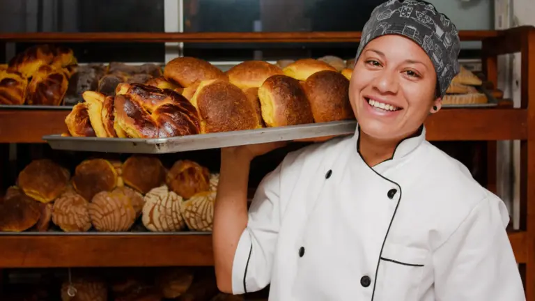 A person in a white chef smock and head wrap holding a tray of golden Mexican pastries on their shoulder and smiling