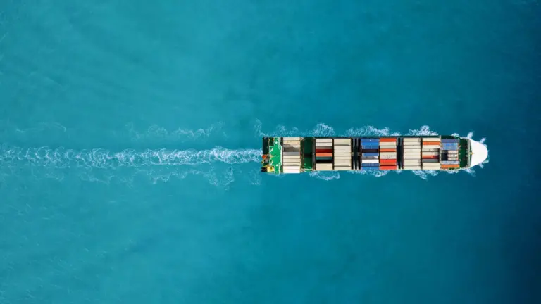 An aerial view of an ocean liner carrying multi-colored shipping containers crossing a turquoise blue ocean