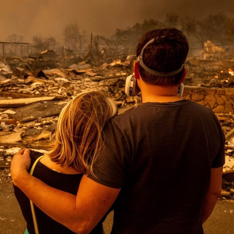 Two people embrace as they look at a burnt home.
