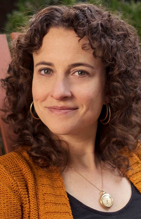 Portrait of Amy Meyerson sitting in chair wearing a brown sweater, black tank top, hoop earrings and a gold chain with a small clock around her neck.