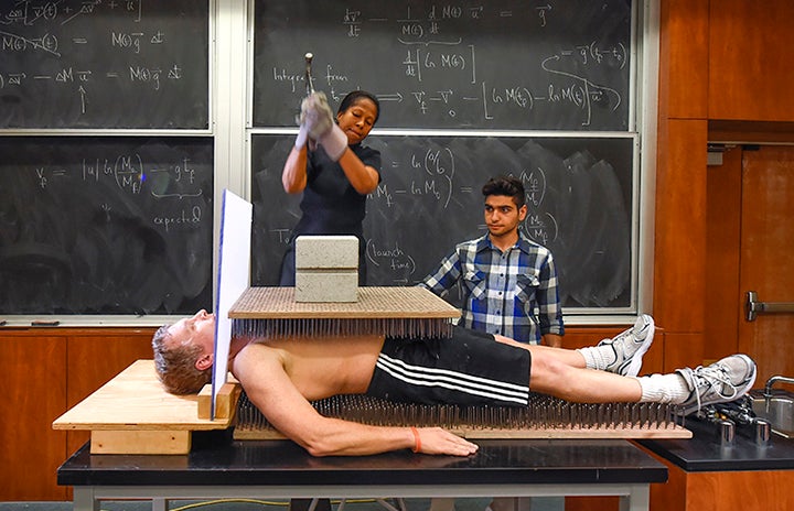 A woman brings down a hammer to break a brick resting on a platfrom of nails that rests on the bare chest of a man laying on a bed of nails as a student looks on.