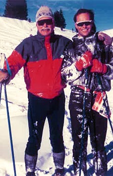 Two men standing next to each other in ski suits on skis. White snow is behind them.