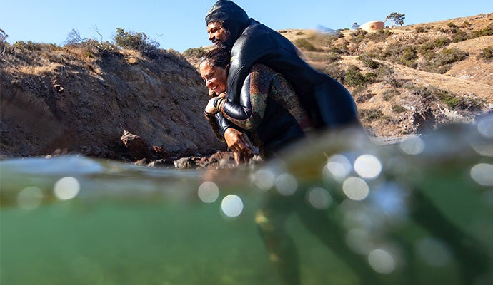 A person in a wet suit carries another person on a wet suit on their back walking through waist deep water.