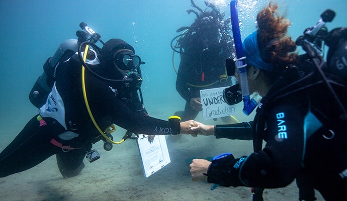 Two scuba divers shake hands underwater, one holds a board and another scuba diver holds up a underwater graduation sign.
