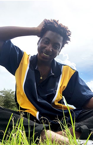 Photo of Felante Charlemagne sitting in the grass with his hand on his head wearing a navy blue, yellow and white shirt and a gold pendant on a chain around his neck