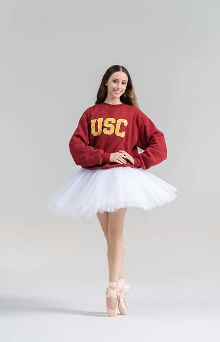 Photo of woman standing en pointe in ballet shoes, wearing a tutu and a cardinal sweatshirt with USC printed on it.