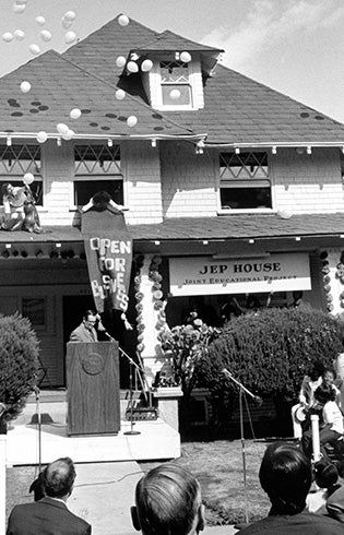 A black and white photo of a building with a sign that says JEP House with balloons being launched in the air, a vertical banner and a podium and mics setup in front with a speaker standing in front of it.