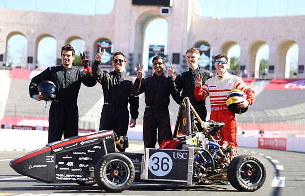 Members of the USC Racing team stand behind a small race car on a track at the L.A. Coliseum..