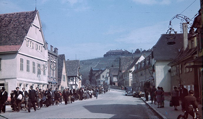 A long line of people carrying their belongings march through a small town.
