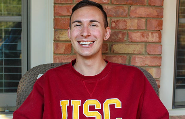 James Fortwengler wears a USC sweatshirt while sitting in a rattan chair against a brick wall and smiling.