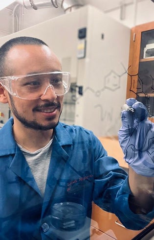 Carlos Navarro wearing safety glasses and gloves writes on a transparent whiteboard in a laboratory.