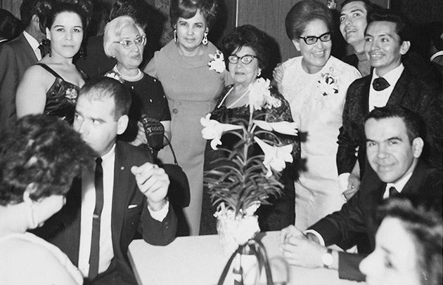 Black and white photo of five women and two men standing behind several people sitting at a table with a tall, flowered plant. 