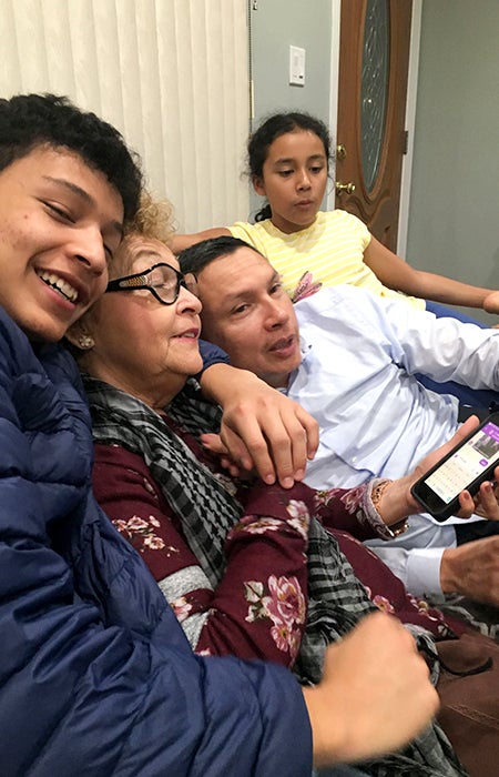 Nelson Henriquez Jr. shares a couch with his family, wrapping his arm around his mother, who holds a cell phone while his father leans on his mother's shoulder and his sister sits next to his father.