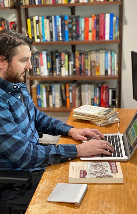 Photo of Ph.D. student Dan Wallace sitting at a desk in an office typing on a computer with several books resting nearby.