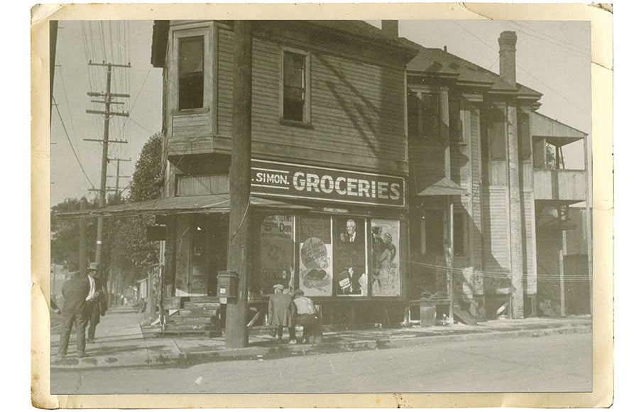 Old sepia-tone photo of the exterior of the Simon Groceries store in East Los Angeles with two men sitting on a bench and pedestrians standing nearby.