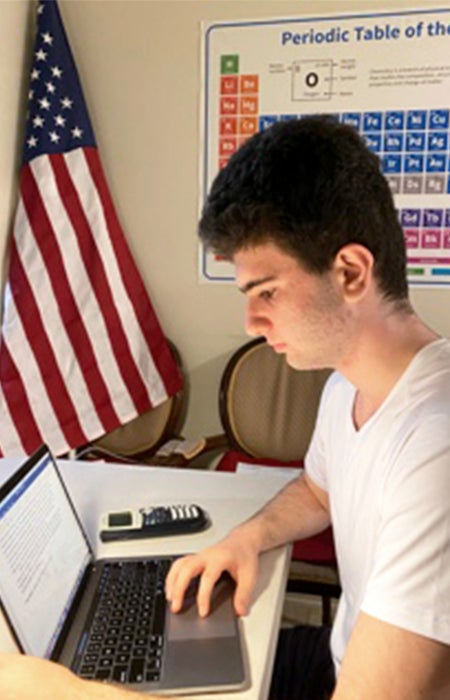 Man seating at laptop. Behind him is an American flag and a periodic table of elements sign.
