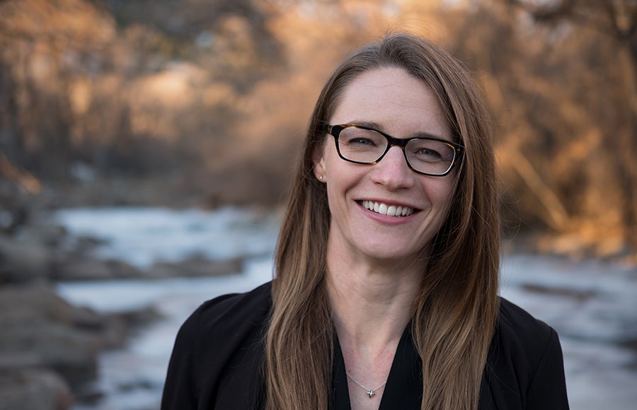 Photo of Alice Baumgartner smiling, wearing glasses and a black top with a stream in the background.