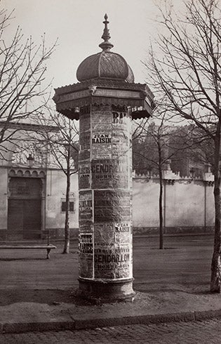 Black-and-white photo of a column topped with an ornate dome and spire and covered in paper advertisements
