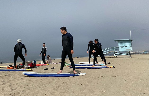 Several people in wetsuits practice standing on a surf board on the beach.