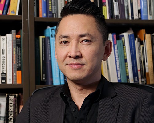 Portrait of Viet Thanh Nguyen wearing a black, collared shirt and dark gray sport coat with shelves full of books behind him