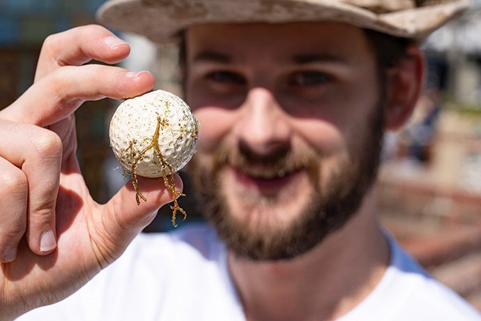 Smiling and bearded Nick Foster wearing a desert camo brimmed hat holds a golf ball with seaweed attached between his thumb and middle finger.