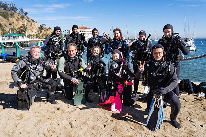 Volunteer divers in scuba suits kneel and stand while holding diving equipment and displaying the "Fight On!" sign with their hands