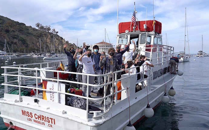 Volunteers wearing face masks wave while standing aboard the white and red boat, Miss Christi