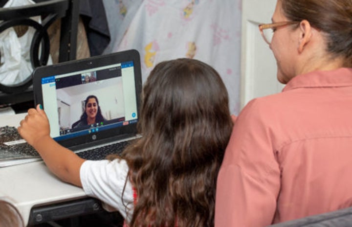 A child sits on her mother's lap and reaches toward a laptop with a college student displayed on the screen.