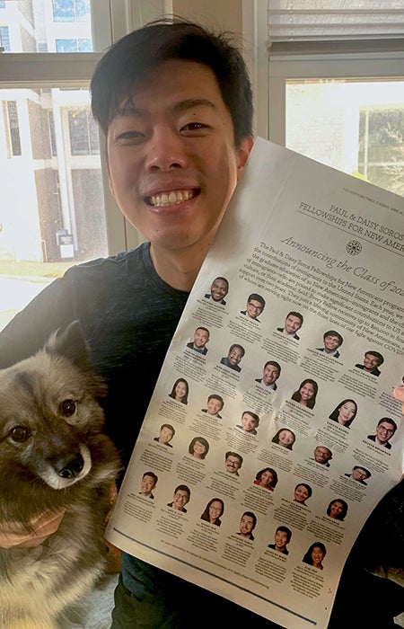 Eric Hoyeon Song smiles while hugging his small dog with his right arm while holding up the 2020 Soros Fellows announcement in his left hand.