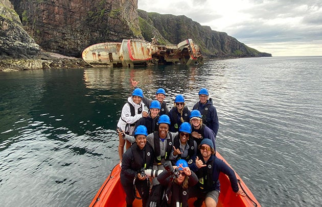 A group poses for a photo wearing helmets in a orange boat in the water with a large vessel capsized on the shore in the background.
