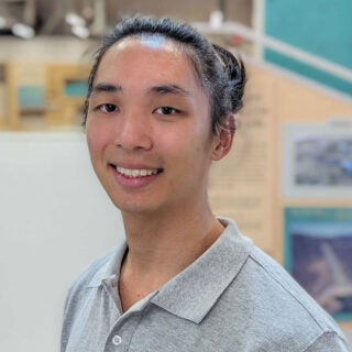 Forrest Lee, an environmental studies alumn who graduated in 2019, wears a gray polo shirt and stands in a warehouse-style space at his employer, the Los Angeles Cleantech Incubator