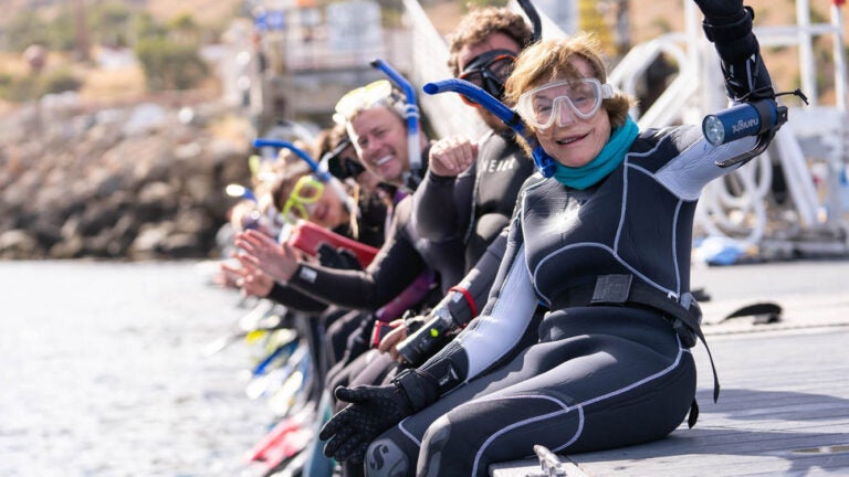 a line of people, wearing wetsuits and snorkels, wave at the camera and smile as they sit on the edge of a dock