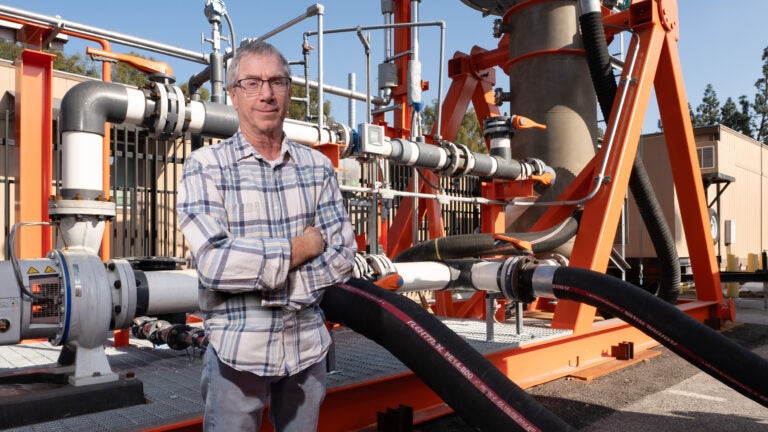 Will Berelson, wearing a flannel shirt and his arms crossed, poses in front of a prototype reactor at USC's University Park Campus