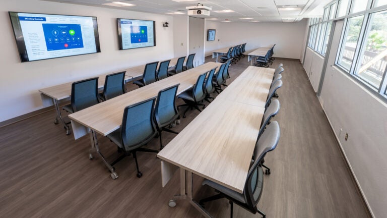 a large boardroom with light-colored tables, blue rolling chairs, and large monitors on the wall