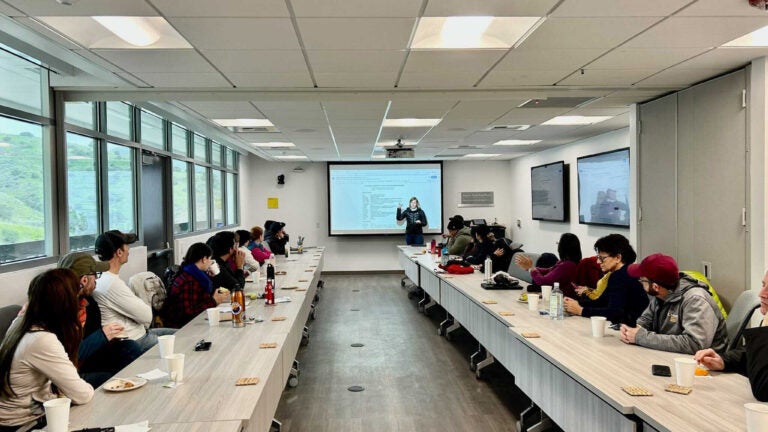 A group of professors, seated at two facing rows of boardroom-style tables, watches a presenter at a large screen at the end of the room.