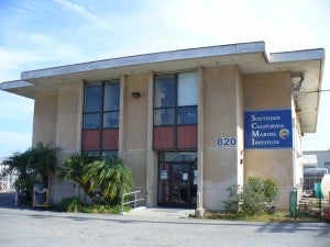 the SCMI building, a two-story, tan stucco building with roof-height windows over the entrance