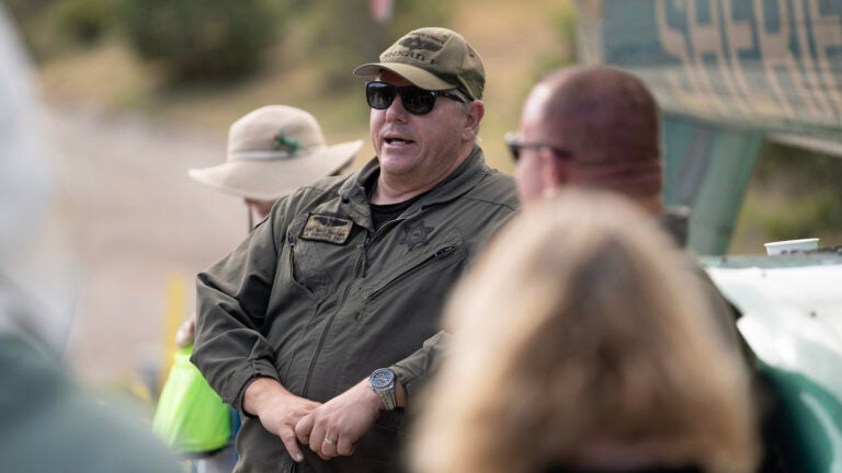 A sheriff leans against their helicopter while talking to a group, hands folded together in front of themself