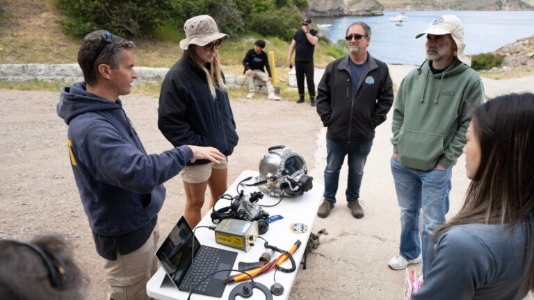 A group of people stand next to a while folding table outside, talking to others about the gadgets present on the table as the harbor sits behind them