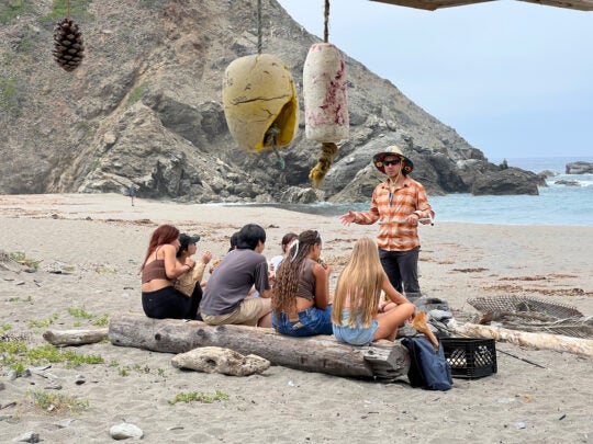 wearing a sun hat and an orange plaid shirt, a professor stands in front of a group of students sitting on driftwood on a beach